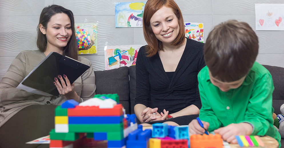 woman with clipboard and another woman sitting watching a small child play with blocks.