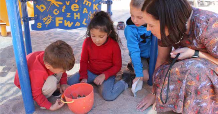 Woman kneeling in a sandbox with three preschool students.