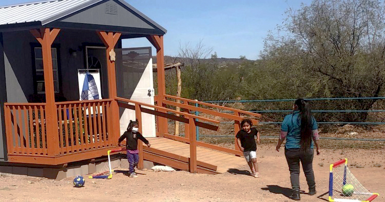 Two young children running outside of their portable classroom as the teacher looks on.