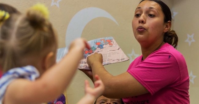 woman in pink tshirt holding up a board book in front of a small child.