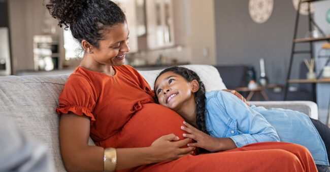 Pregnant mother on couch in orange dress with young girl hugging the mother's stomach