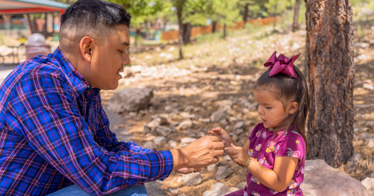 Dad in blue, long sleeve shirt showing little girl in purple something outside near a tree. Making nature accessible