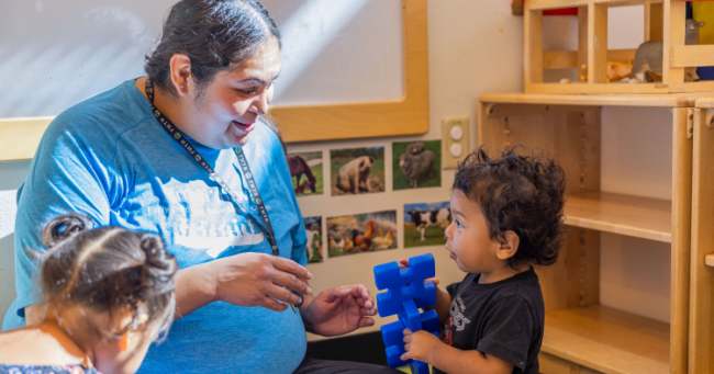 Child care worker in blue shirt talking to a little boy with a blue toy in his hand. early care and education workforce